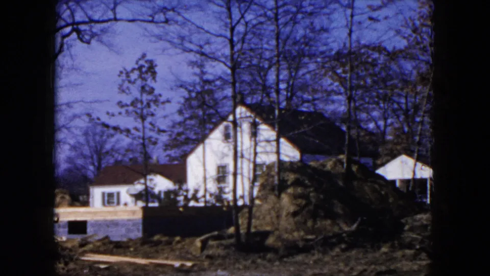 1954 Hicksville frame with a white house behind a foundation wall and large dirt mound, framed by leafless trees and 8mm borders.
