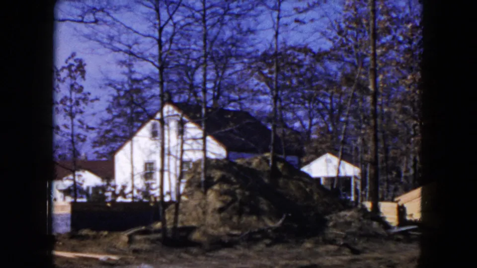 1954 Hicksville home-movie frame showing a suburban lot with a white house, dirt pile, small shed, bare trees, and building materials.