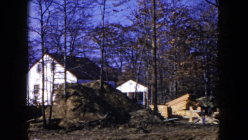 1954 Hicksville frame showing a residential construction lot with a white house, mound of earth, bare trees, small shed, and stacked lumber.