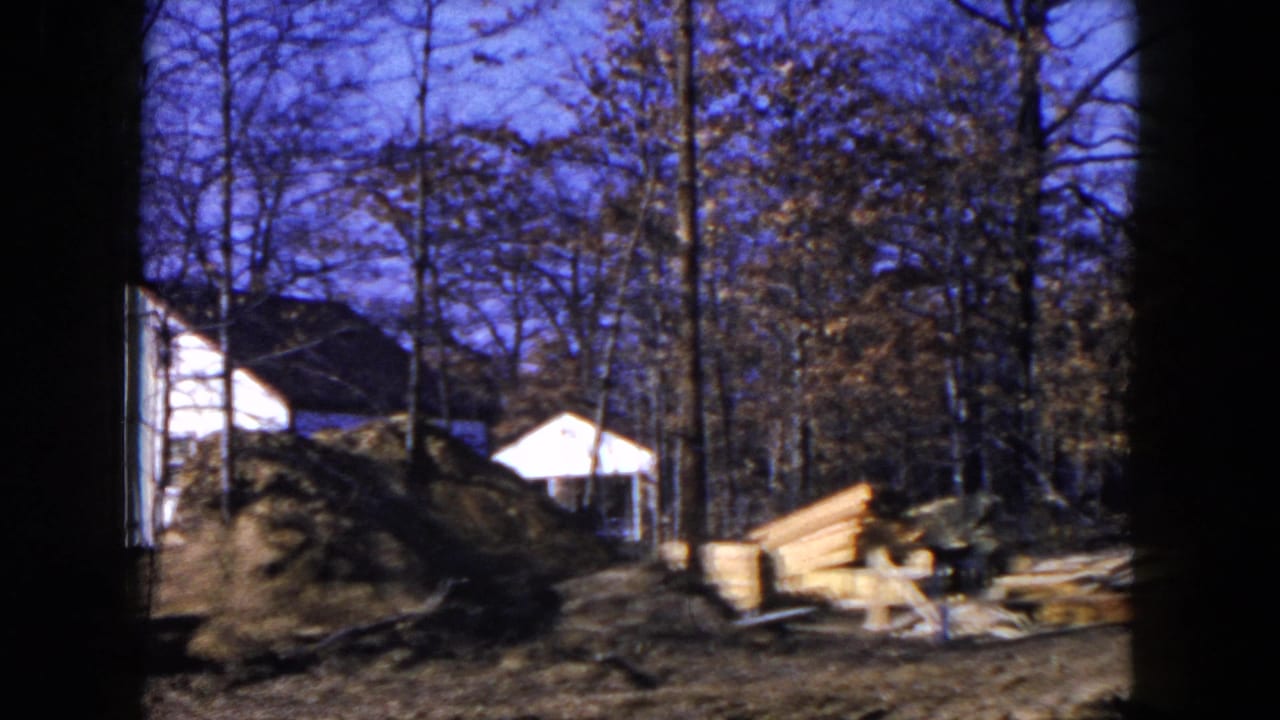 1954 Hicksville home-movie frame showing a wooded residential construction site with stacked lumber, muddy ground and a small white structure beyond the trees.