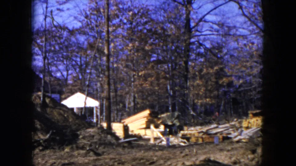 1954 Hicksville frame showing construction materials in a wooded residential lot, with lumber piles and a small white structure partly visible behind trees.