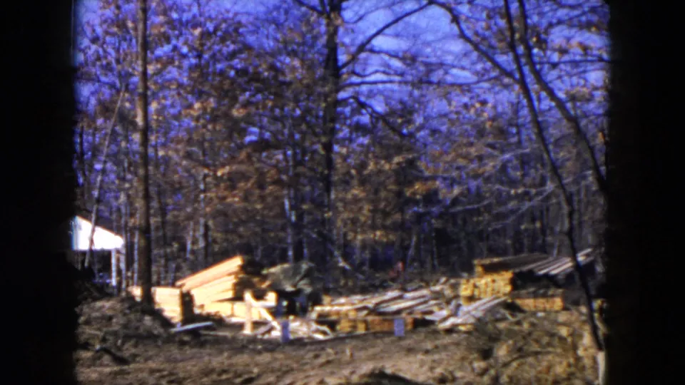 1954 Hicksville home-movie frame of a muddy wooded construction area, with stacked lumber, scattered boards and a small white building at the left edge.