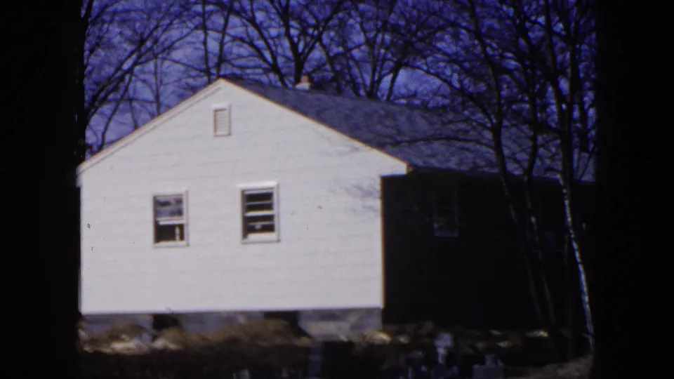 1954 Hicksville frame showing a white house exterior on a raised foundation, surrounded by bare trees with a dark side structure attached or nearby.