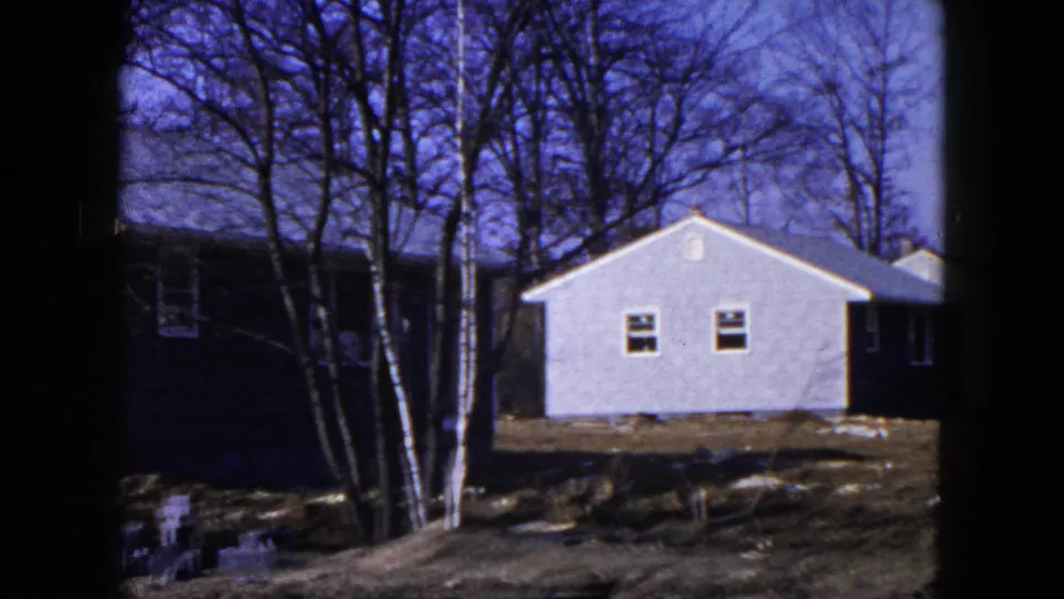 1954 Hicksville frame showing a small pale gabled house beyond uneven ground, with bare trees and adjacent houses nearby.