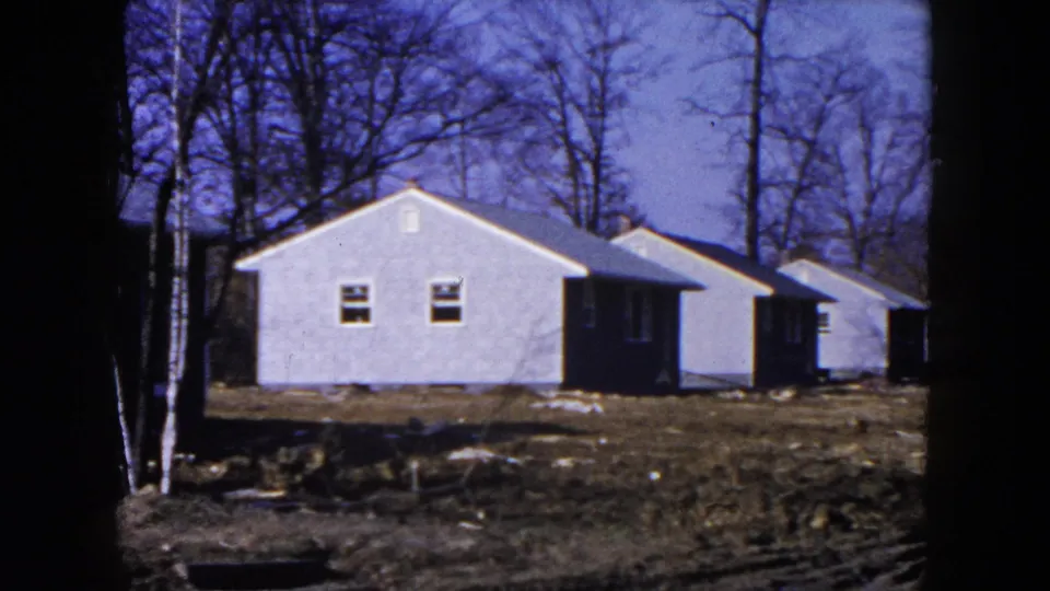 1954 Hicksville frame of a pale house on an unfinished lot, with bare trees, neighboring houses, and visible 8mm frame borders.