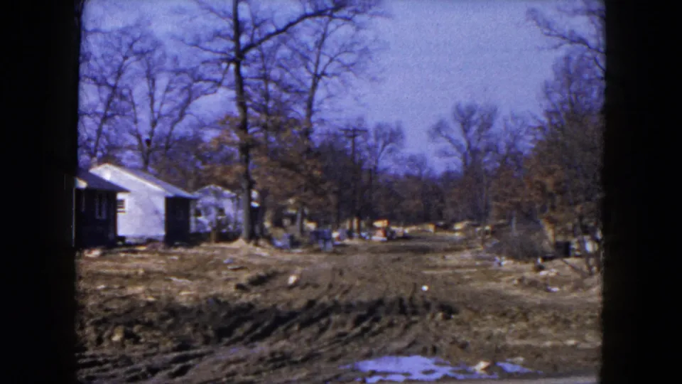1954 Hicksville, New York frame showing the edge of white houses beside muddy ruts, snow patches and leafless trees.