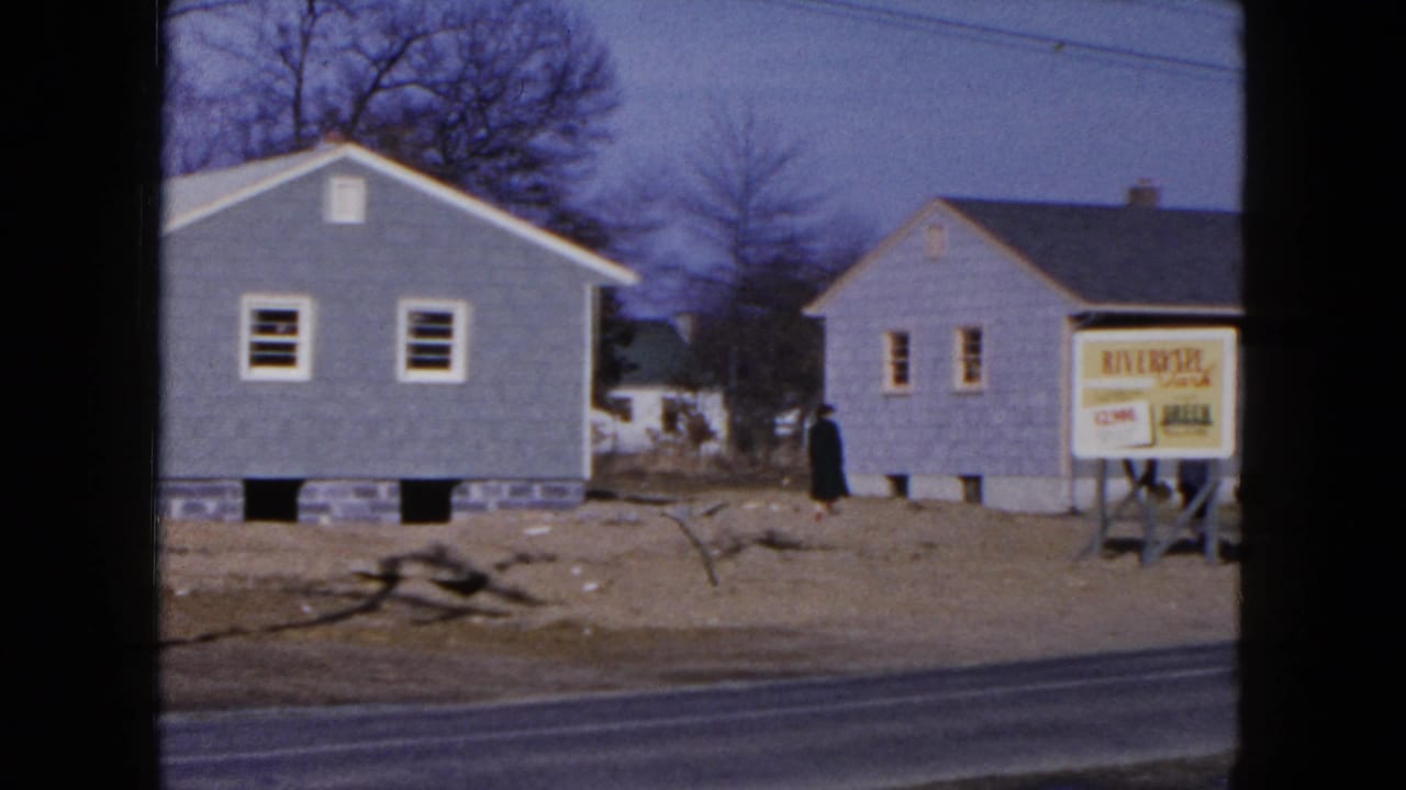 Two small gray houses beside a road with a real estate sign and a person walking nearby.