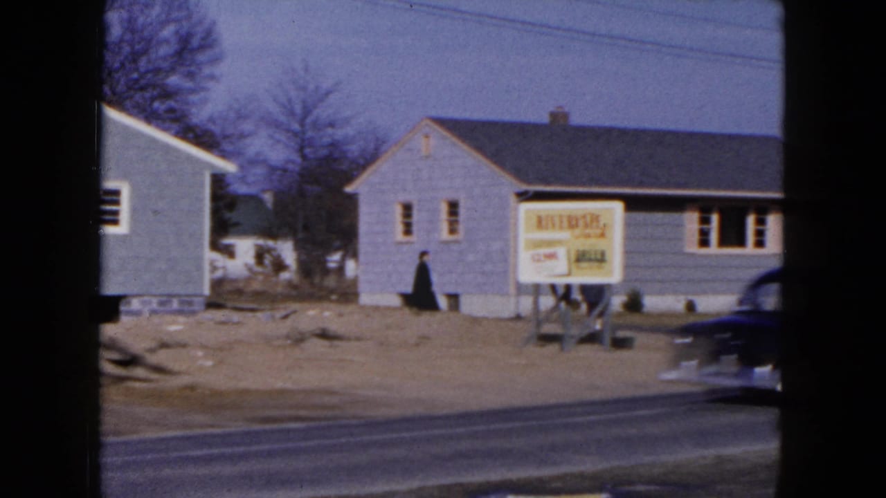 1954 Hicksville frame with gray suburban houses, the Riverside sign, bare trees, and a dark mid-century car passing on the road.