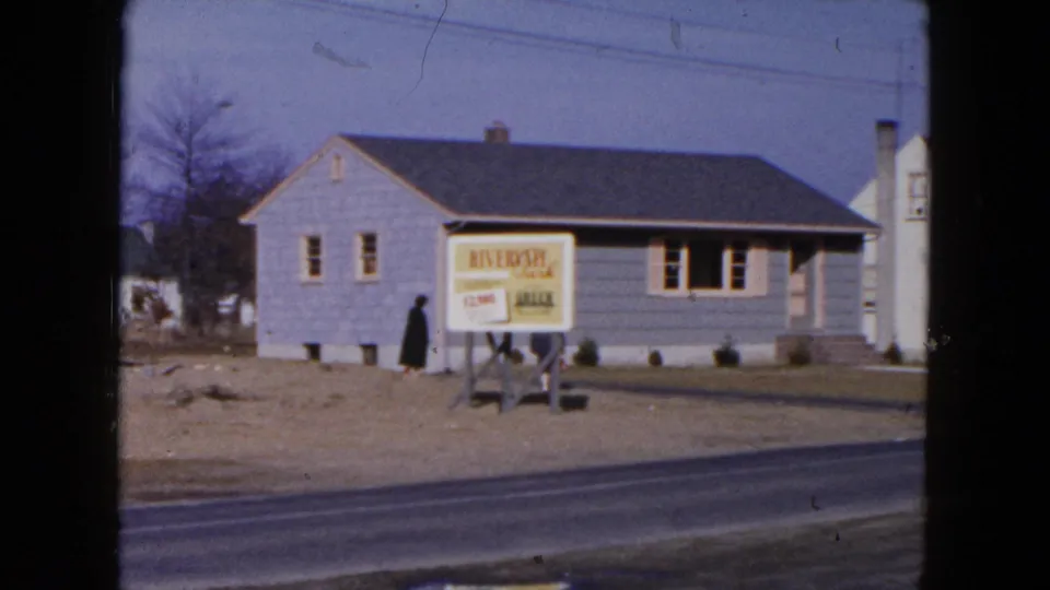 1954 Hicksville frame showing a dark-clothed pedestrian near the Riverside sign, with a gray house, bare trees, and roadside foreground.