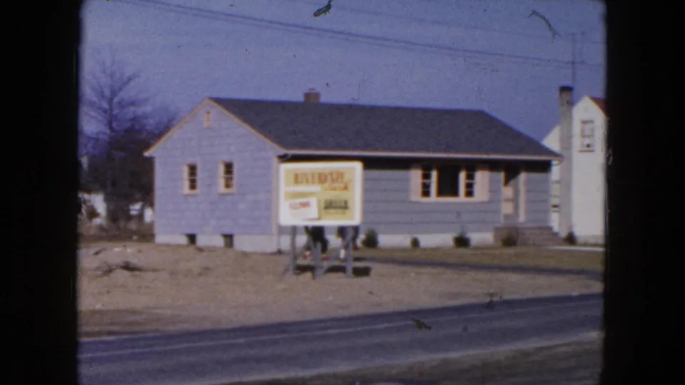 1954 Hicksville home-movie frame with a gray suburban house, Riverside real estate sign, paved road, bare trees, and a white structure at right.