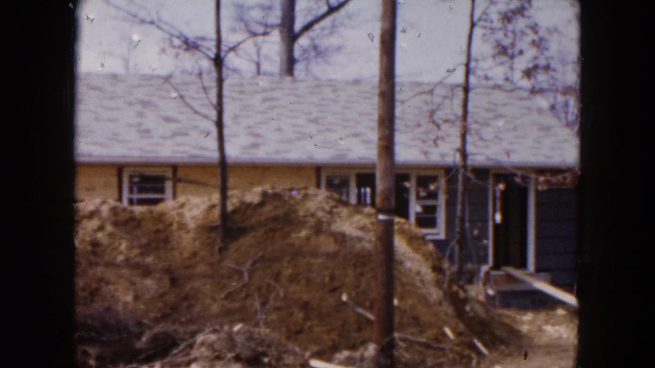 Blue and yellow house under construction behind a dirt mound and bare trees.