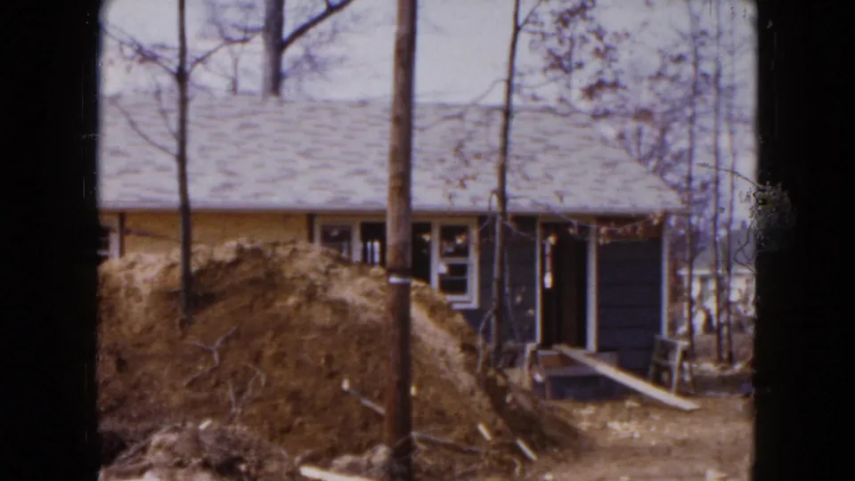 1954 Hicksville frame of the house exterior during construction, with a dirt pile, trees, doorway, and plank access visible.