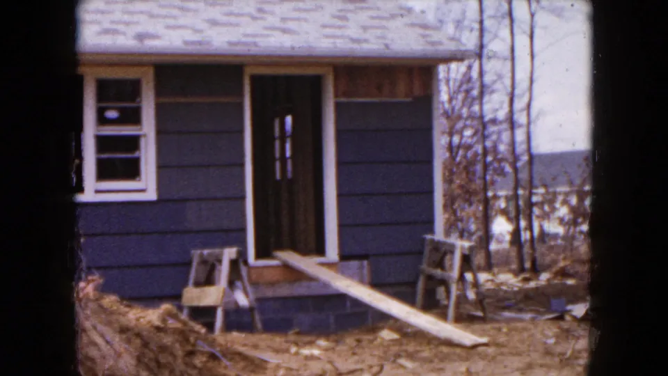 1954 Hicksville home-movie frame showing the house entrance, blue siding, plank walkway, sawhorses, and wooded surroundings.