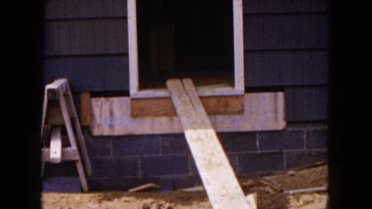 1954 Hicksville home-movie frame showing an unfinished house exterior with dark siding, an open window, wood planks, and a sawhorse.