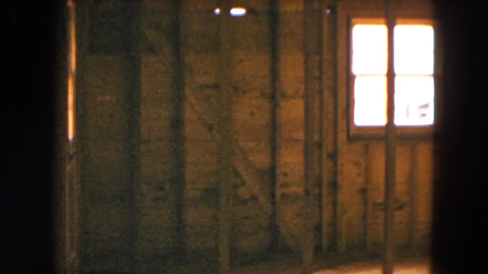 Inside the Hicksville house under construction, a rough wooden wall and small bright window are visible in dim 8mm light.