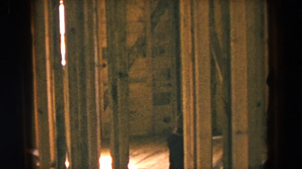 Low-light 8mm frame inside the Hicksville construction site, showing vertical framing, a narrow bright opening, and unfinished flooring.