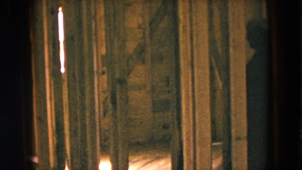 1954 Hicksville home-movie frame showing a dim unfinished room with exposed wooden studs and warm light in the background.