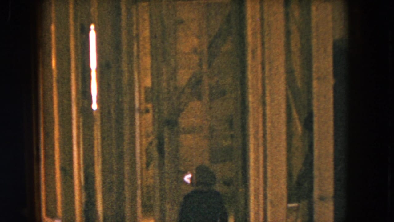 Small dark-clothed figure near exposed framing in a dim room.