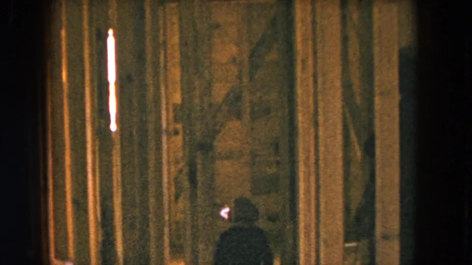 A small figure in dark clothing stands near exposed framing inside the dim 1954 Hicksville residential interior.