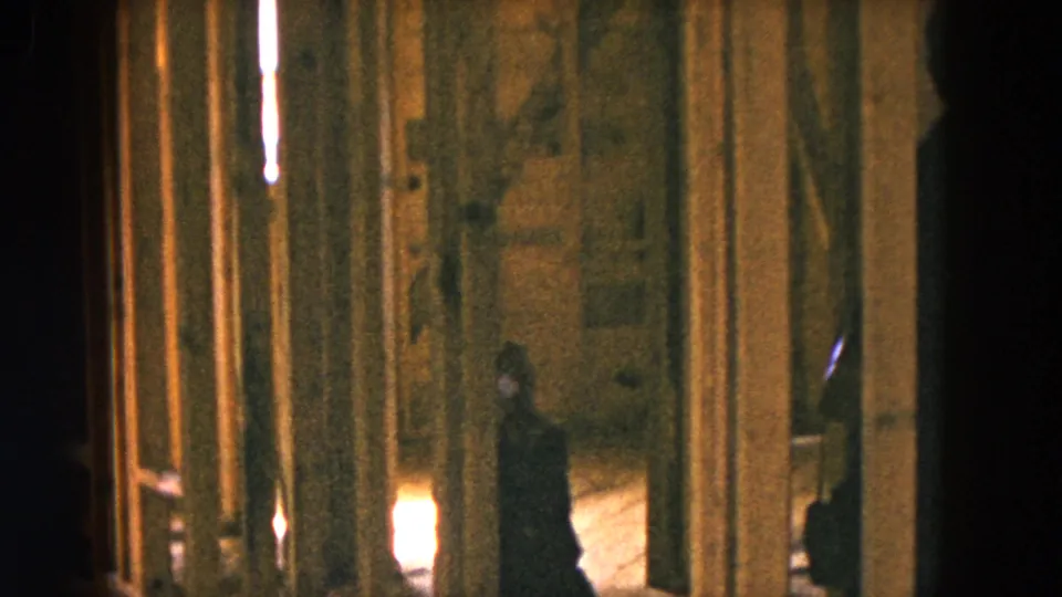 The child stands in the dim unfinished room while a dark adult figure appears near the right edge of the 1954 home-movie frame.