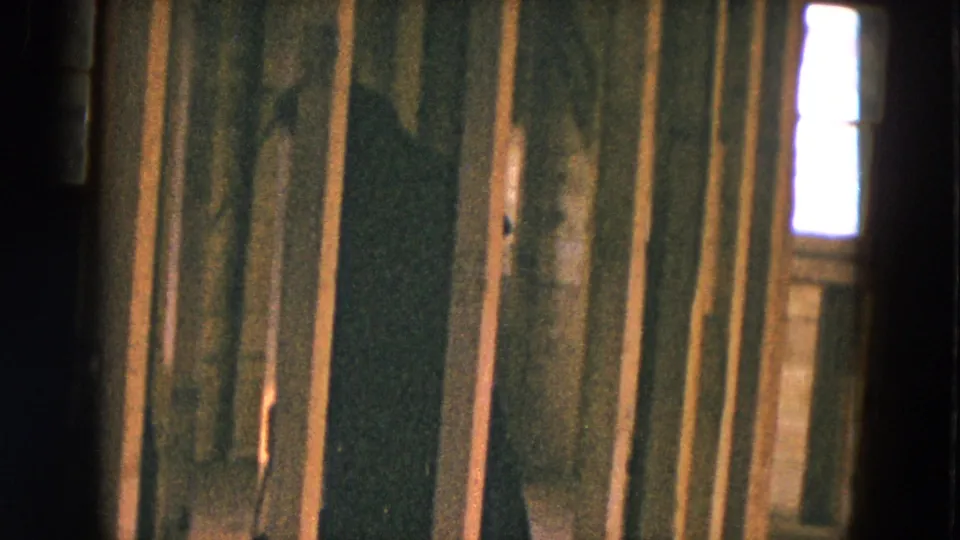 Handheld 8mm view of an unfinished house interior, with rough lumber, a dark doorway, and bright window light beyond.