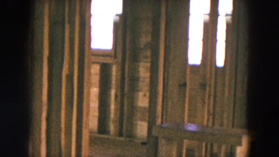 1954 home construction frame showing exposed studs, bright narrow windows, and a low horizontal piece of lumber inside the unfinished house.