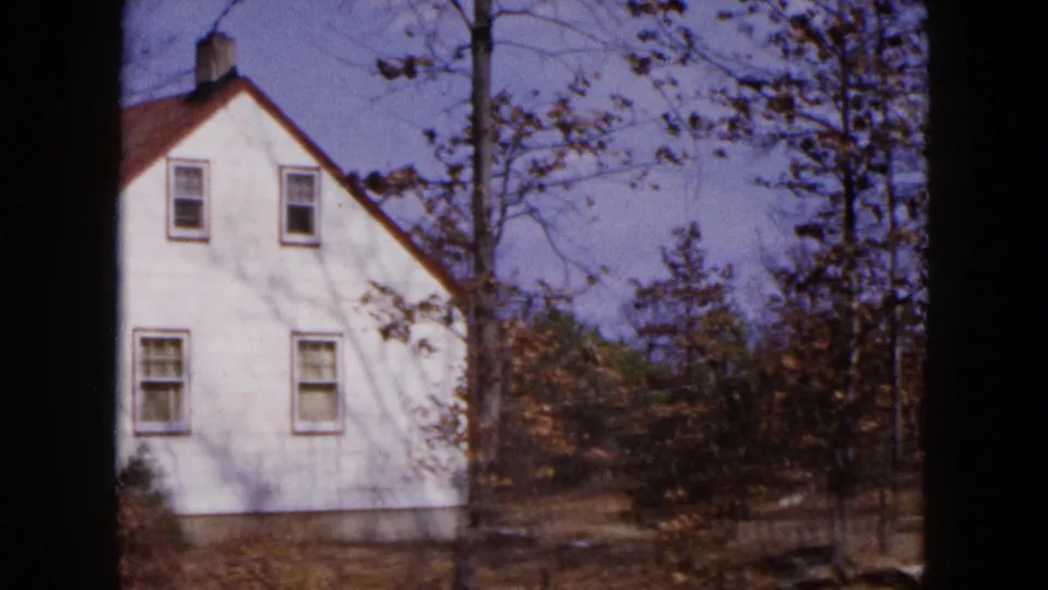 1954 8mm frame of the house side in Hicksville, with windows, a bare tree, brush, and wintery yard.