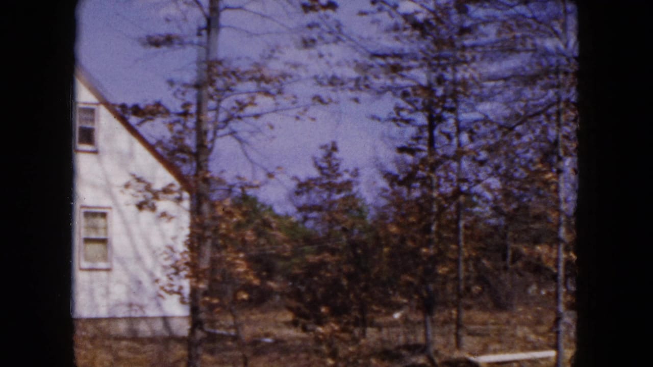 1954 Hicksville home-movie frame showing the edge of a white house behind leafless trees and brush.
