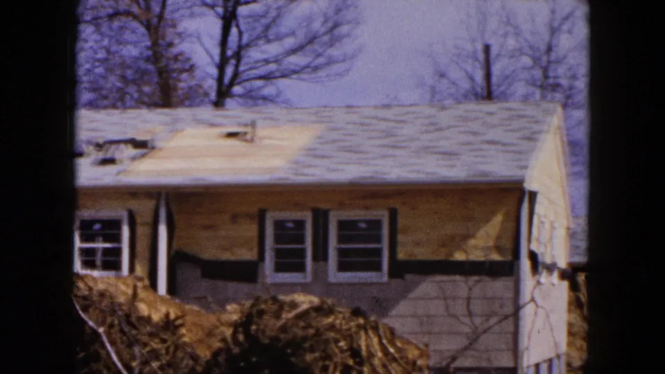 A side portion of the Hicksville house shows unfinished exterior walls, a nearly covered roof plane, window openings, and construction soil in front.