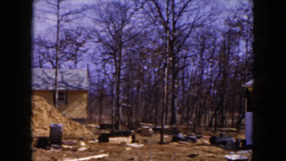 1954 Hicksville view across a debris-strewn yard with a dirt pile, bare trees, and partial damaged house exterior.