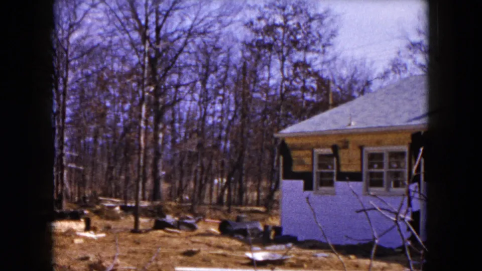 1954 Hicksville home-movie frame showing a house edge, wooded yard, debris, and disturbed ground after storm damage.