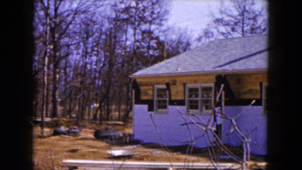 1954 Hicksville home-movie frame showing a single-story house under exterior renovation, with purple lower siding, exposed wood above, and yard debris.