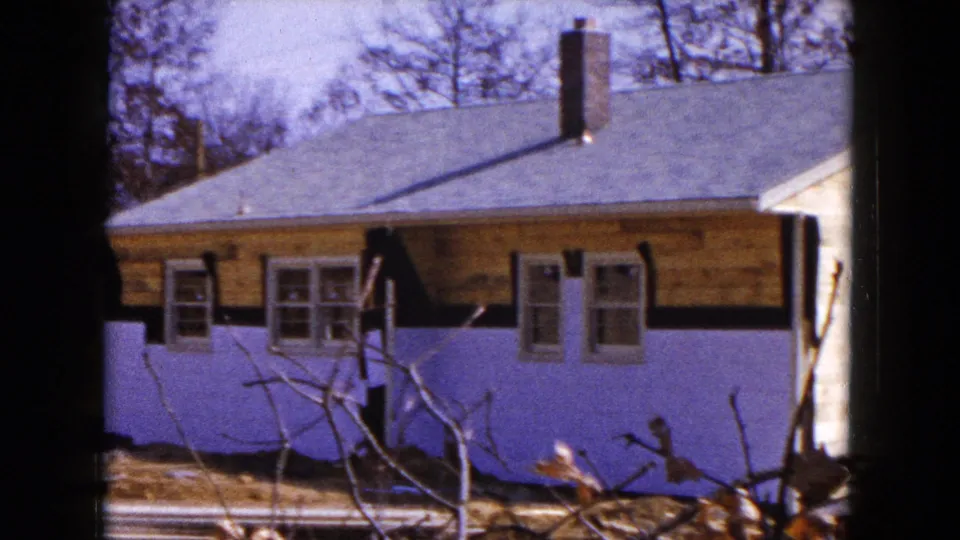 1954 Hicksville frame showing the front facade of a small house with dark shutters, unfinished upper siding, purple lower walls, and winter-bare trees.