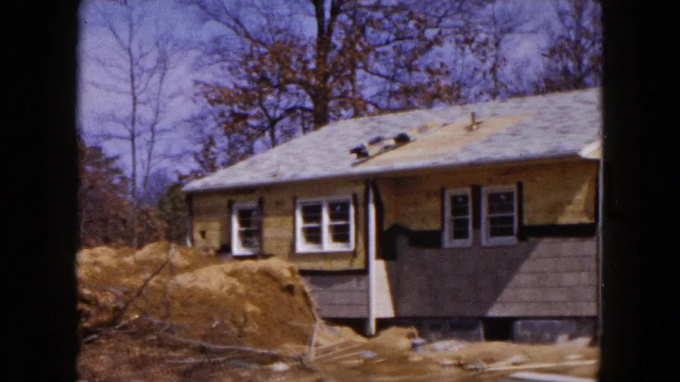 1954 Hicksville frame showing a side view of the renovated house, with purple lower siding, exposed wood, bare trees, and construction debris in the yard.