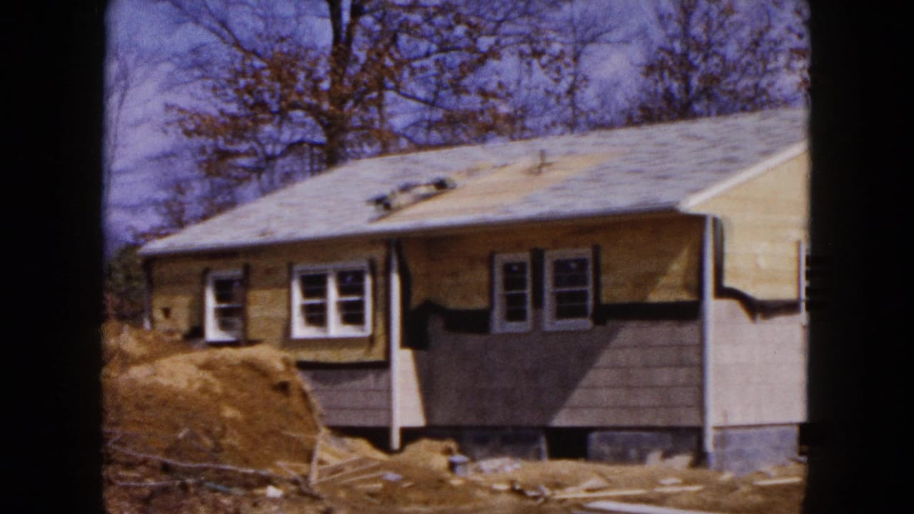 1954 Hicksville home-movie frame showing a single-story house under construction with exposed siding, roof vents, dirt piles, and autumn trees.