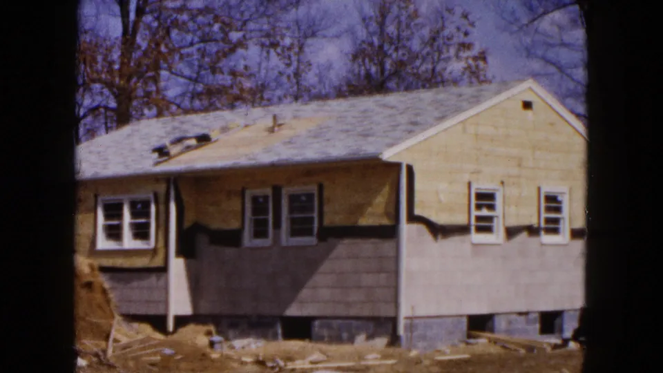 1954 Hicksville frame of the house exterior during construction, with gray siding sections, roof vents, installed windows, and bare trees in back.