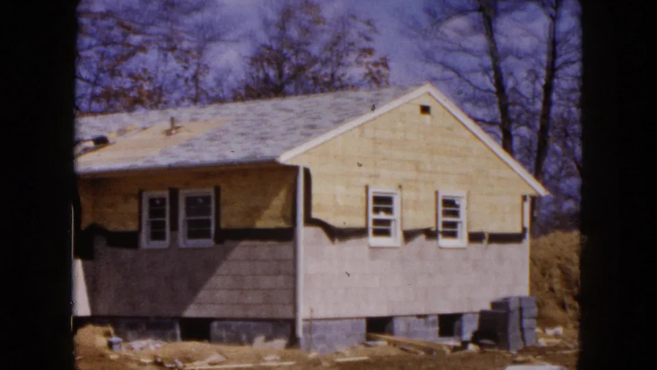 1954 Hicksville home-movie frame showing the gable end of the unfinished house, with windows, roofline, exposed foundation openings, and materials nearby.