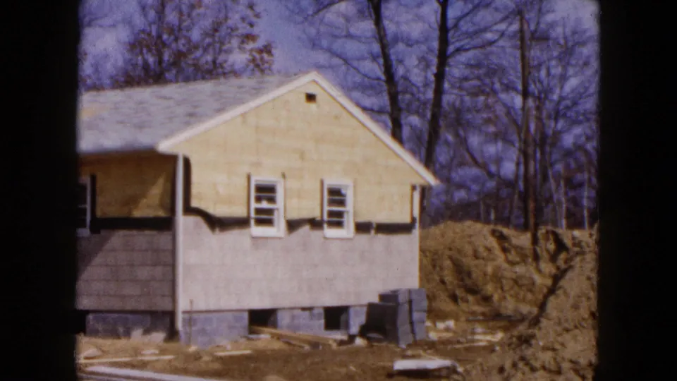 1954 Hicksville frame showing the gable side of the house under construction, with small windows, exposed foundation, stacked blocks, and autumn trees.