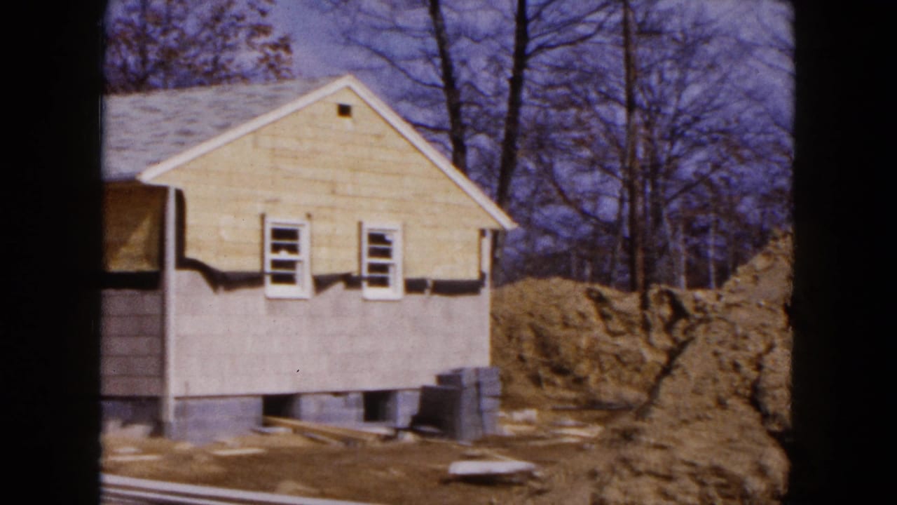 1954 Hicksville home-movie frame showing a yellow-sided house under construction with bare trees and piled earth nearby.
