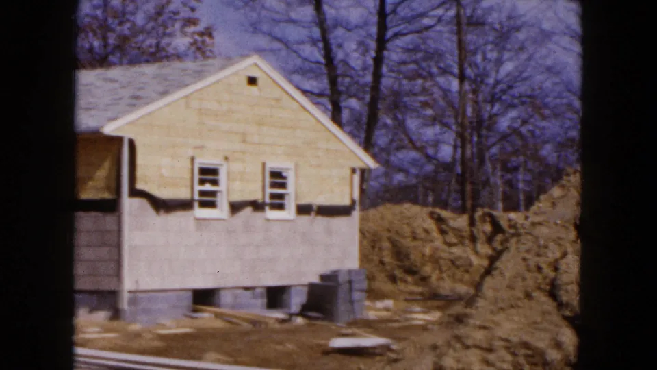 1954 Hicksville home-movie frame showing a yellow-sided house under construction with bare trees and piled earth nearby.