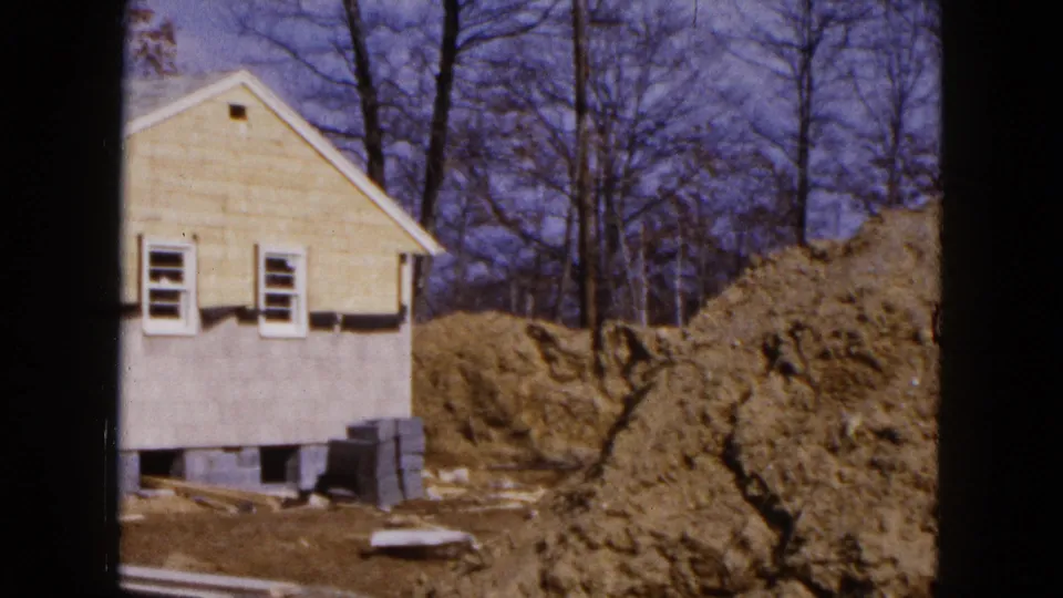 1954 Hicksville frame of a partially built house, exposed foundation, piled soil, and leafless trees in the background.