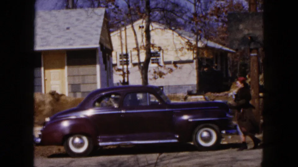 1954 Hicksville street view with a maroon sedan parked near a house and a woman in dark clothing walking past.
