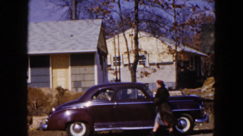 1954 Hicksville frame showing a woman beside a parked maroon sedan, with a house and construction earth behind her.