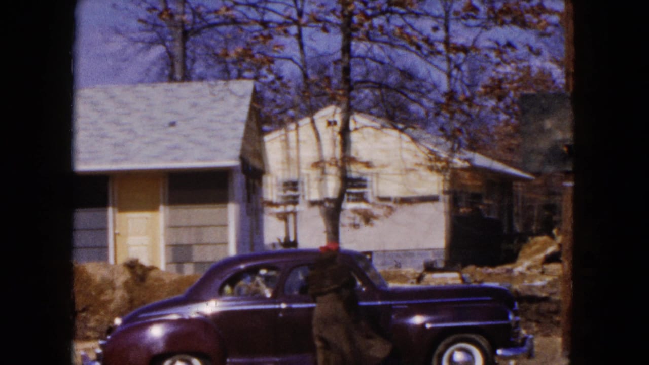 Dark sedan parked near a light house as a person in a red hat stands beside the car.