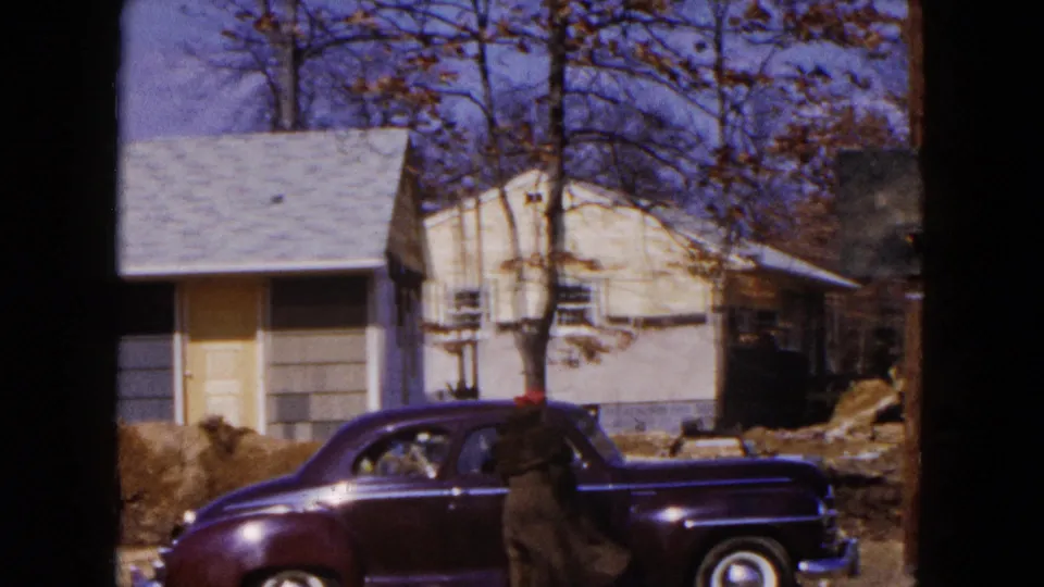 1954 Hicksville home-movie frame showing a dark sedan, a person in a red hat, a light house, bare trees, and disturbed ground.