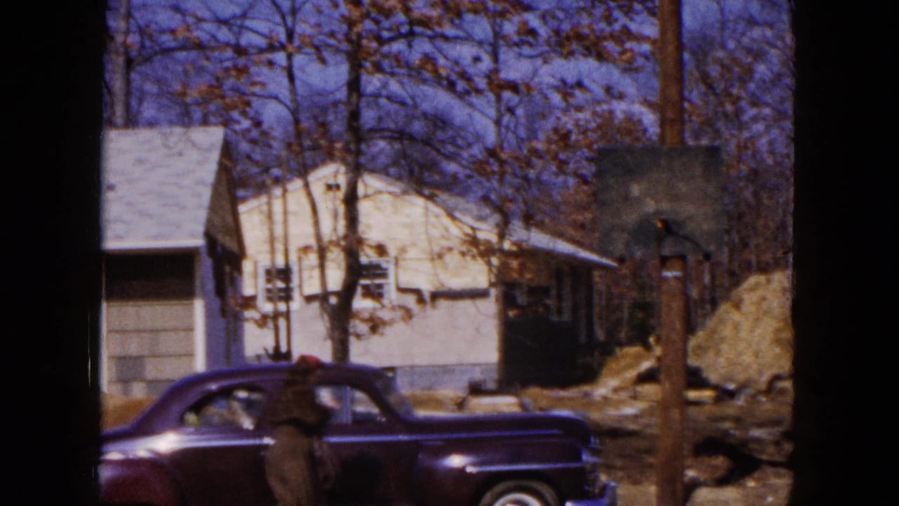 Dark sedan and standing person pass across the lower frame in front of a house and utility pole.