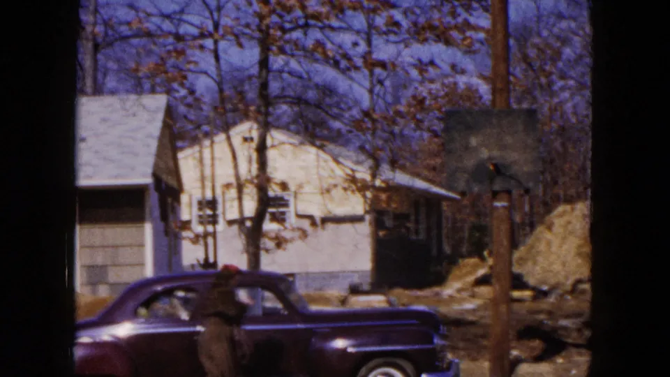 1954 Hicksville frame with a dark sedan at curbside, a standing figure, a light house, bare trees, and a wooden utility pole.