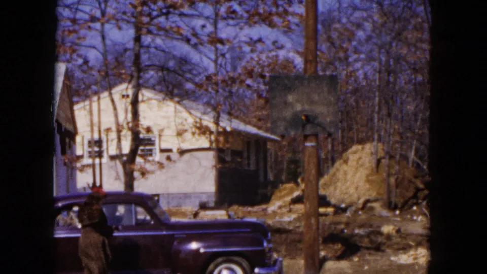 1954 Hicksville home-movie still showing the sedan partly cropped, a person nearby, a small house, pole, trees, and renovation debris.
