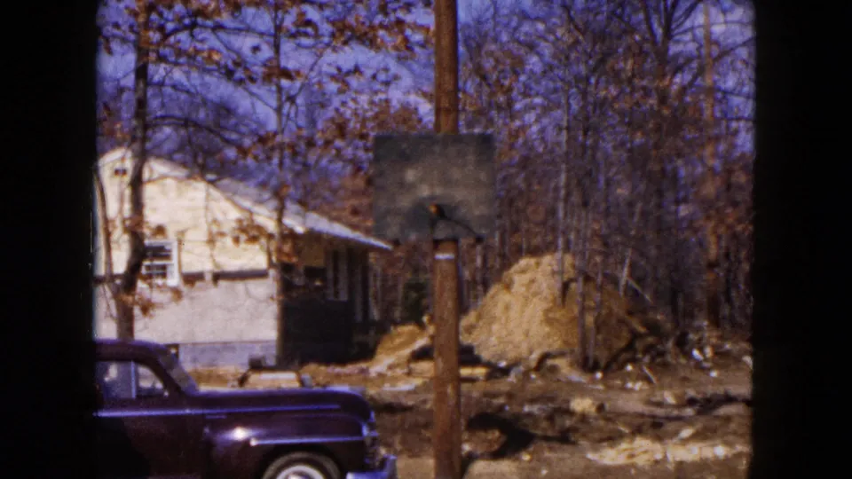 1954 Hicksville frame with the dark sedan partly visible below a small house, wooden pole, bare trees, and uneven construction ground.