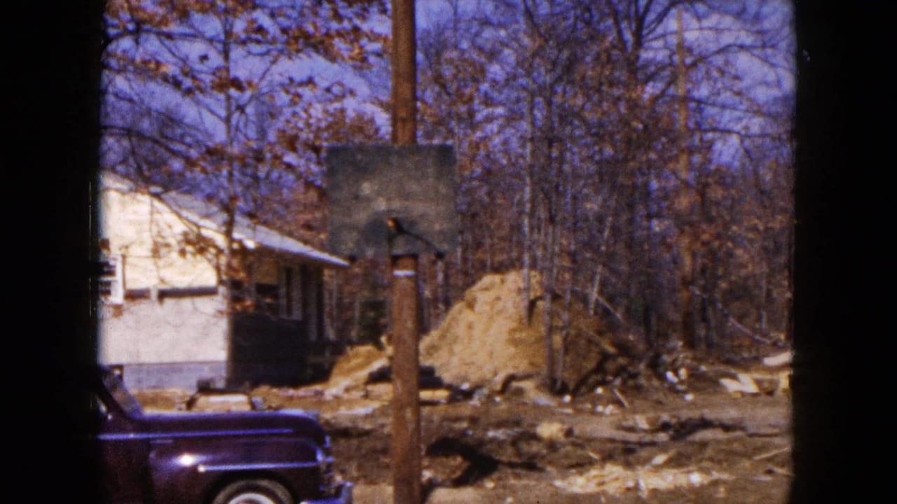 1954 Hicksville home-movie frame showing a house under construction with dirt piles, bare trees, a utility pole, and a dark sedan.